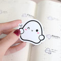 Hand holding a cute cartoon ghost sticker over an open notebook with more stickers on the page.