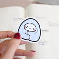 Hand holding a sticker with a cartoon ghost design on a notebook page.
