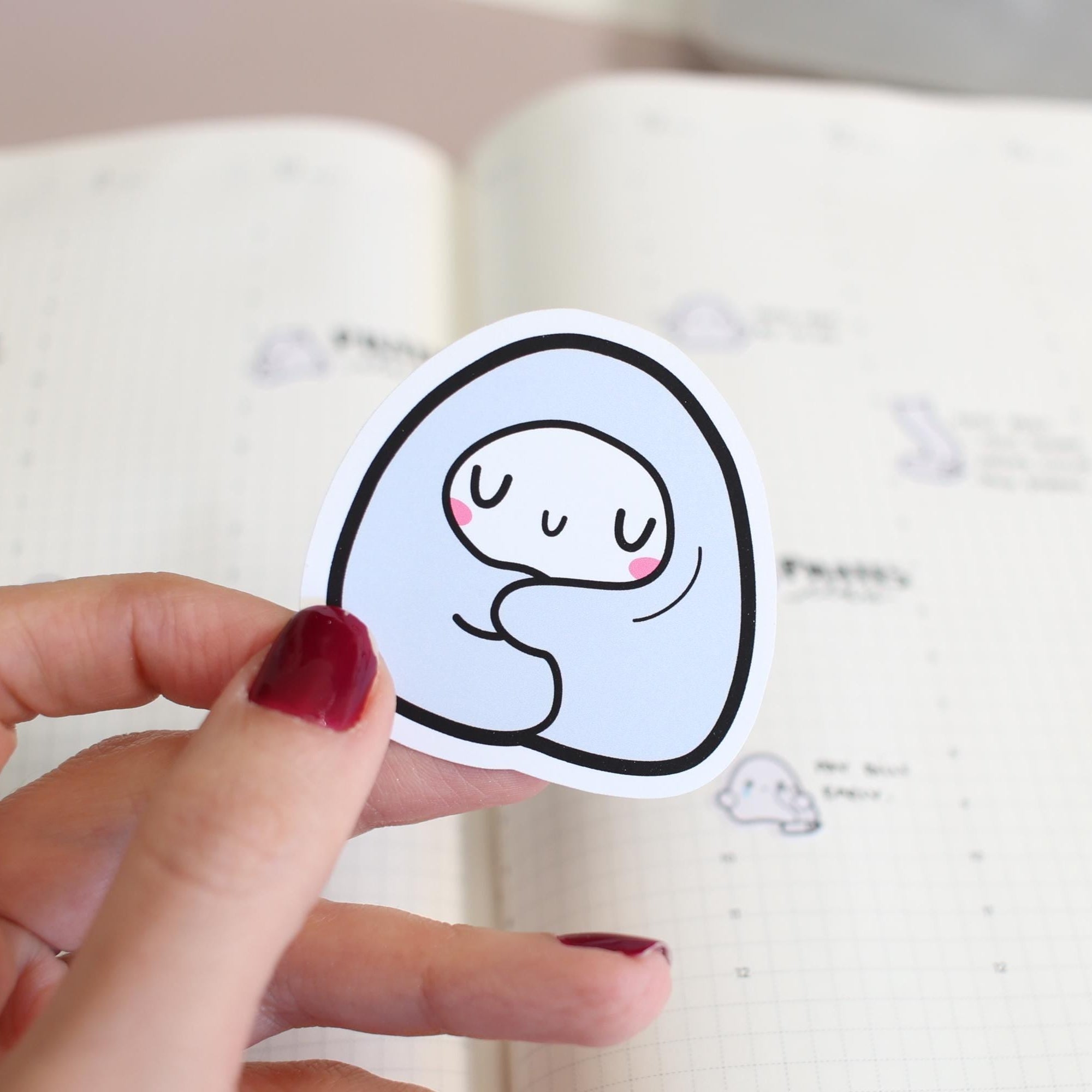 Hand holding a sticker with a cartoon ghost design on a notebook page.