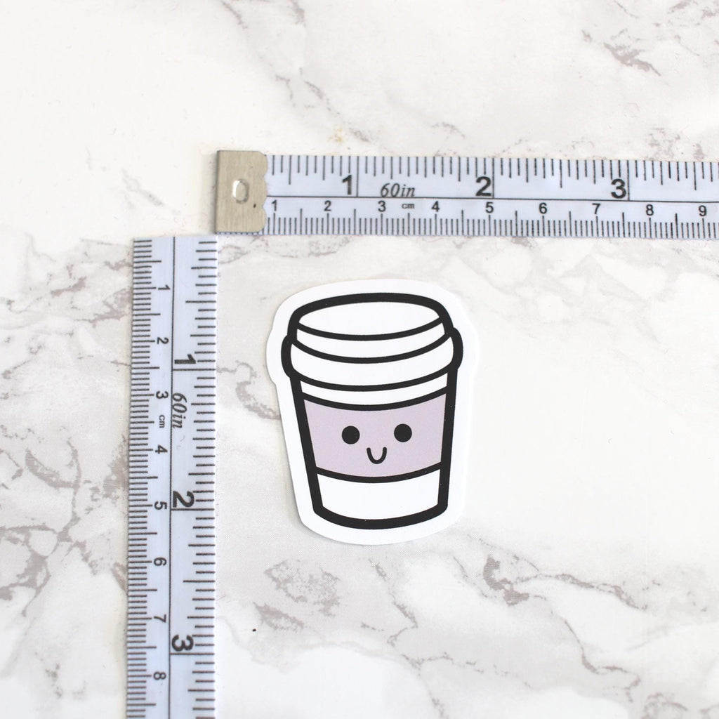Sticker of a coffee cup with a smiley face next to a ruler on a marble surface