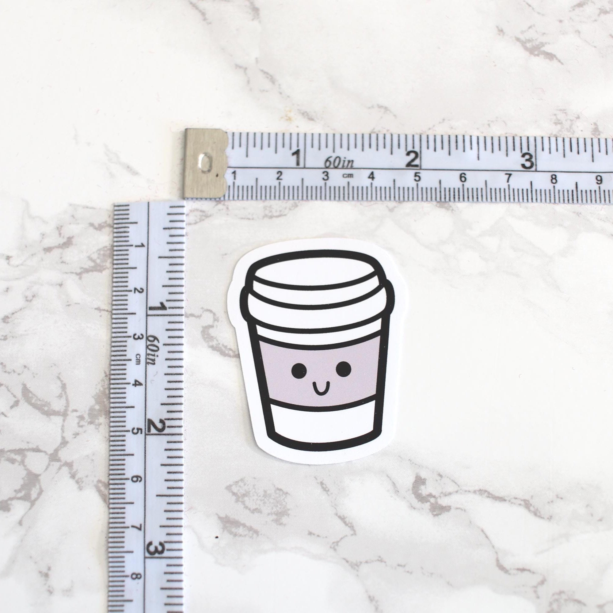 Sticker of a coffee cup with a smiley face next to a ruler on a marble surface