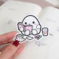 Hand holding a cute cartoon sticker of a character reading a book with a cup and snacks, over an open notebook.