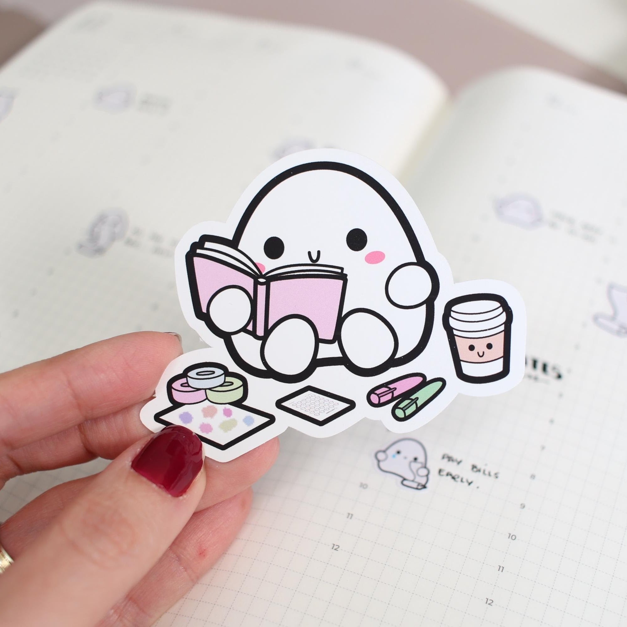 Hand holding a cute cartoon sticker of a character reading a book with a cup and snacks, over an open notebook.