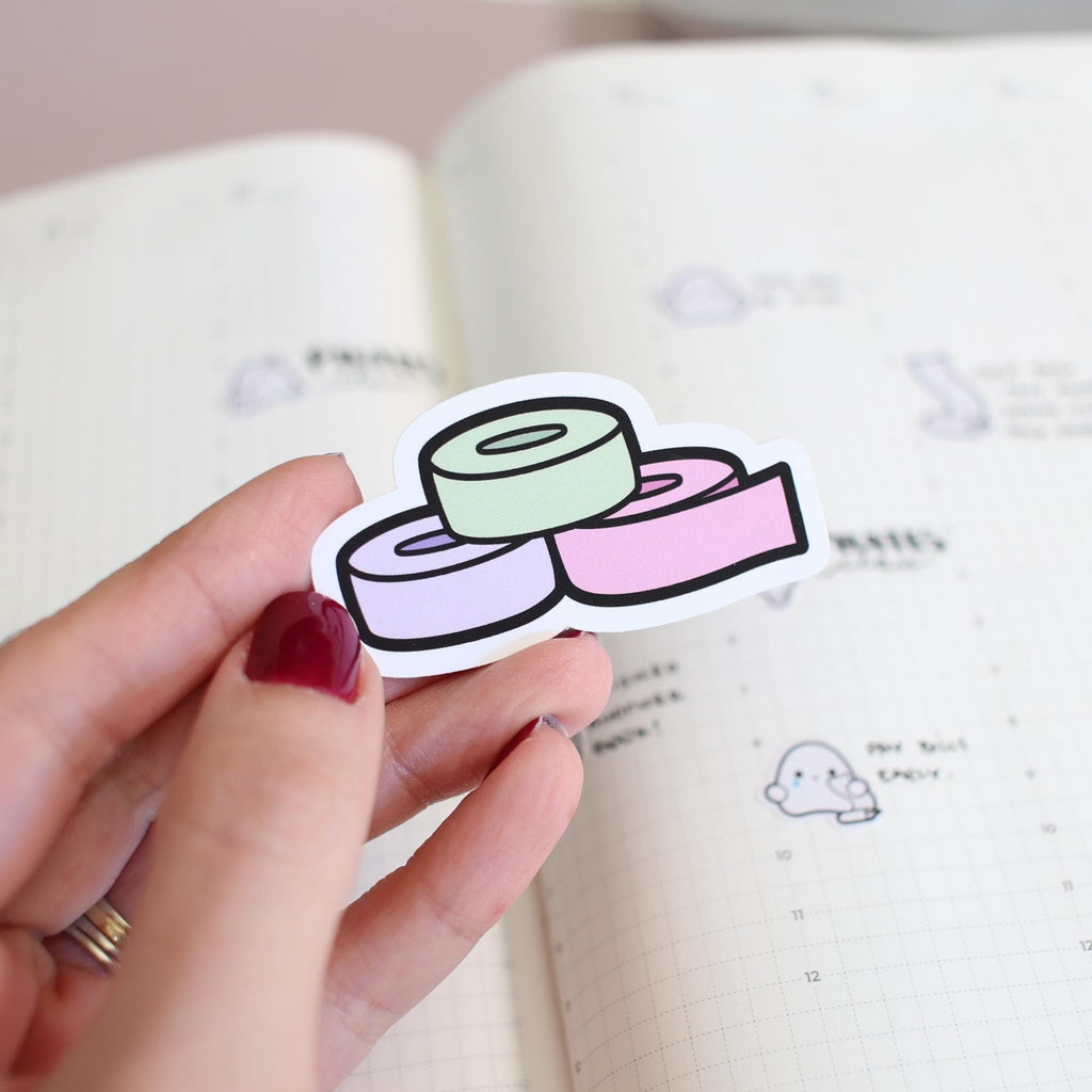 Sticker of three pastel green, purple, and pink rolls with black outlines, held by a hand with red polished fingernails and a gold ring above an open planner displaying grid lines and a faint cartoon ghost sticker