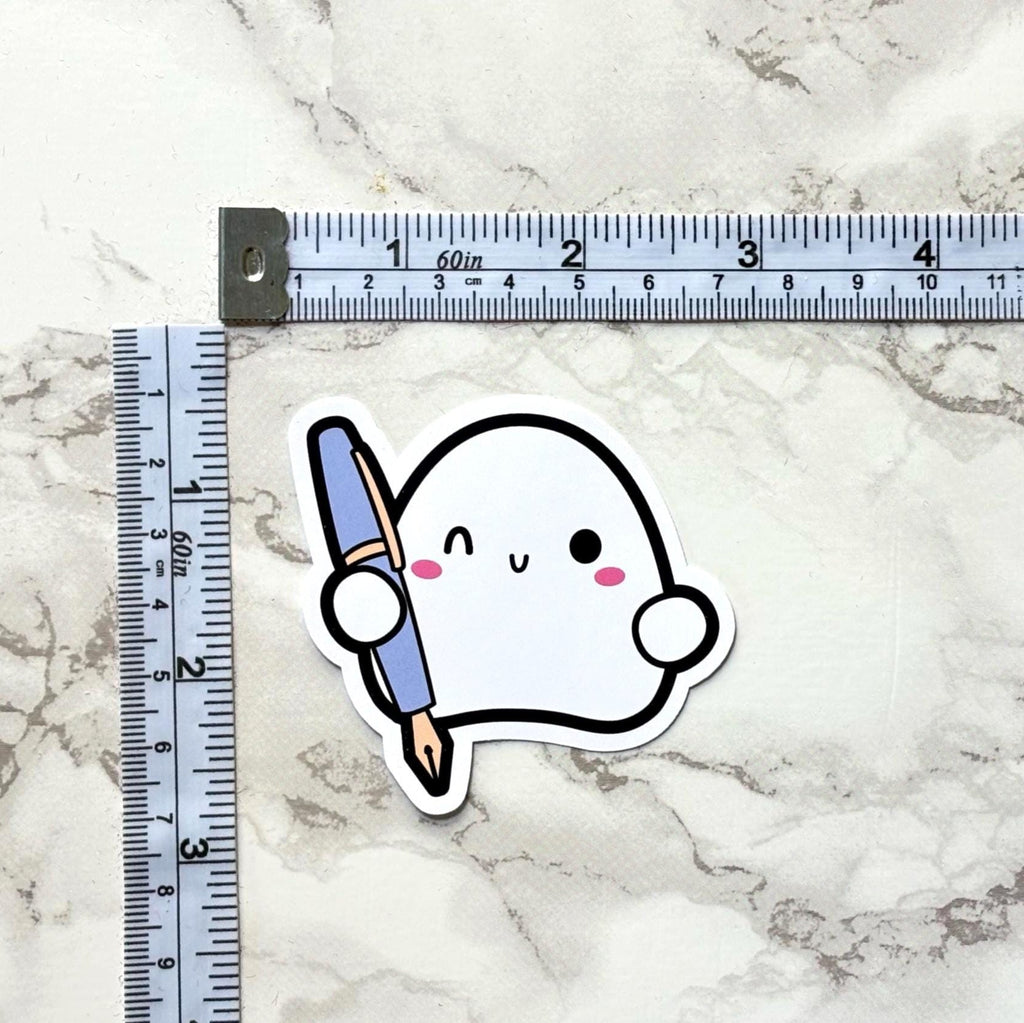 Sticker of a cute ghost holding a paintbrush next to a ruler on a marble surface