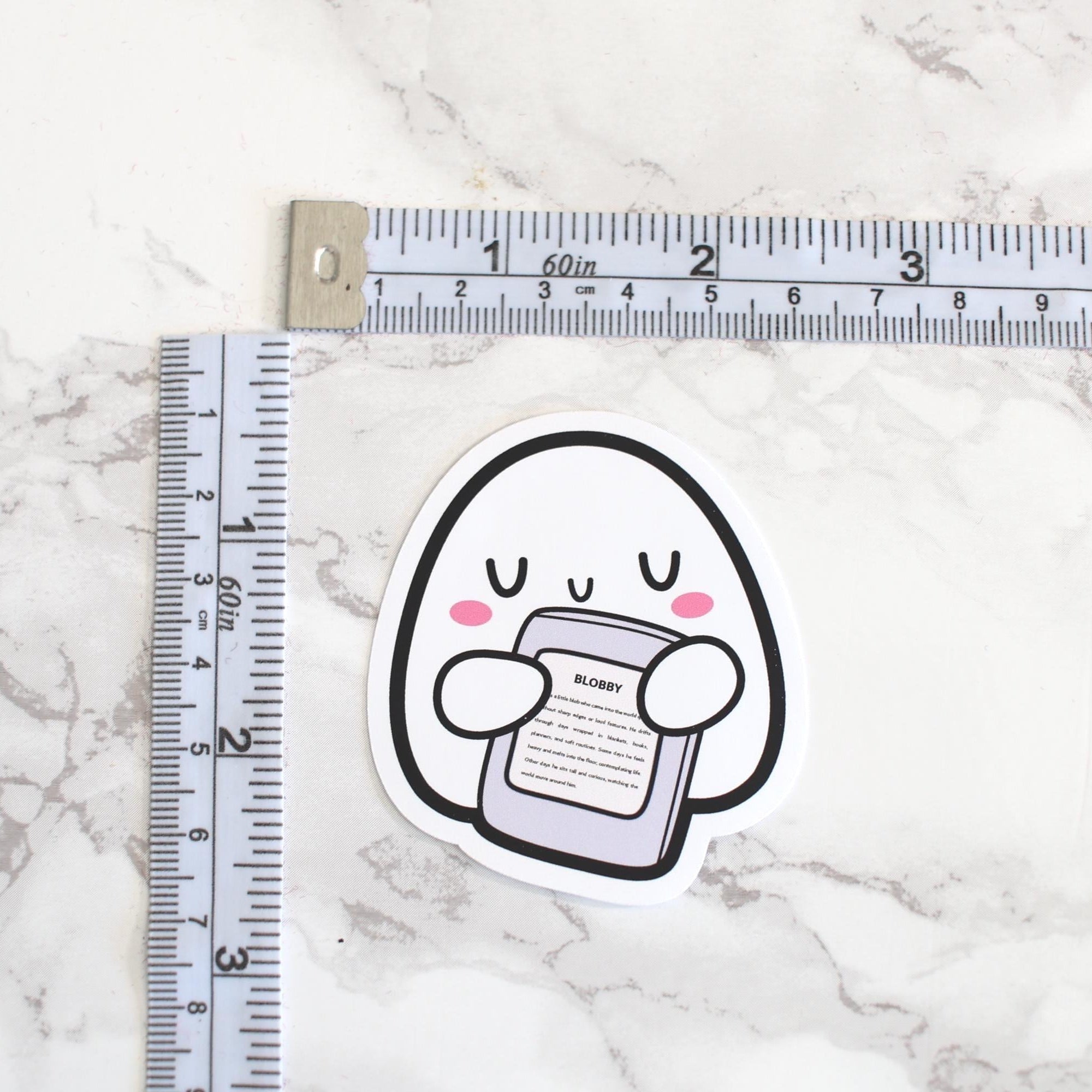 Sticker of a cartoon character holding a phone next to a ruler on a marble surface