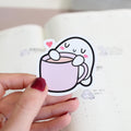 Cartoon sticker of a cup with a face held by a hand on a notebook background
