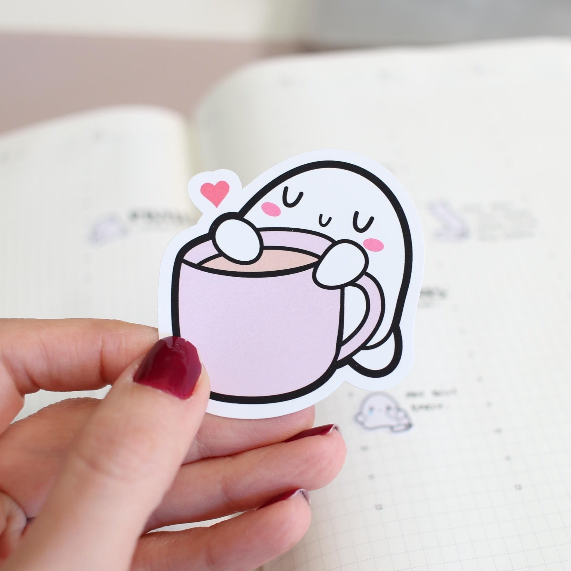 Cartoon sticker of a cup with a face held by a hand on a notebook background