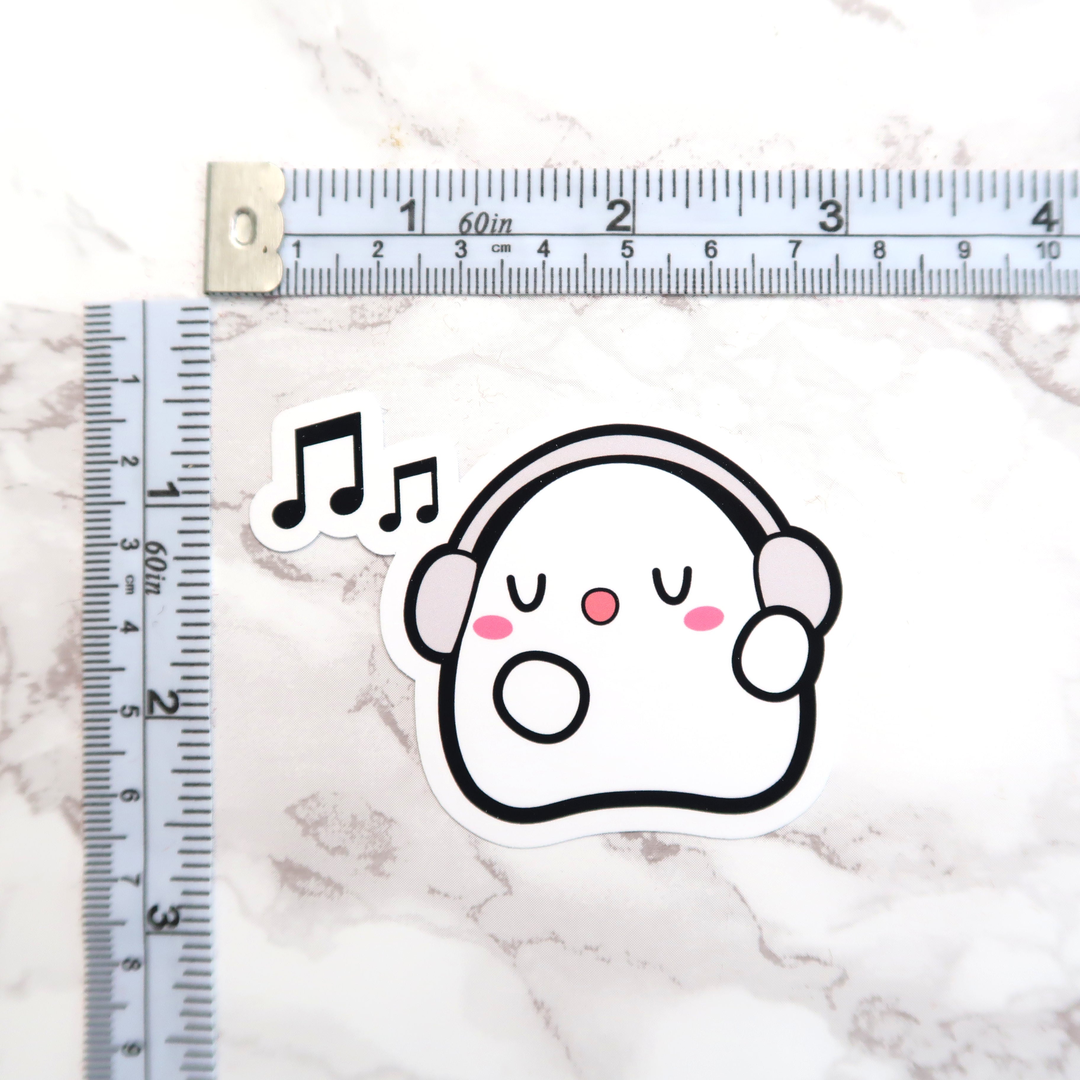 Sticker of a white cartoon ghost wearing light grey headphones with pink cheeks and nose, and two black music notes floating above its head, resting on a white marble background next to horizontal and vertical measuring tapes