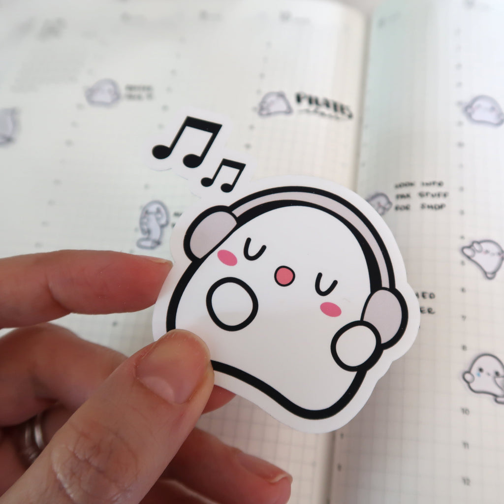 Sticker of a white cartoon ghost wearing grey headphones, singing happily with pink cheeks and black musical notes floating above, held by a hand against a grid-lined planner