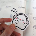 Sticker of a white cartoon ghost wearing grey headphones, singing happily with pink cheeks and black musical notes floating above, held by a hand against a grid-lined planner