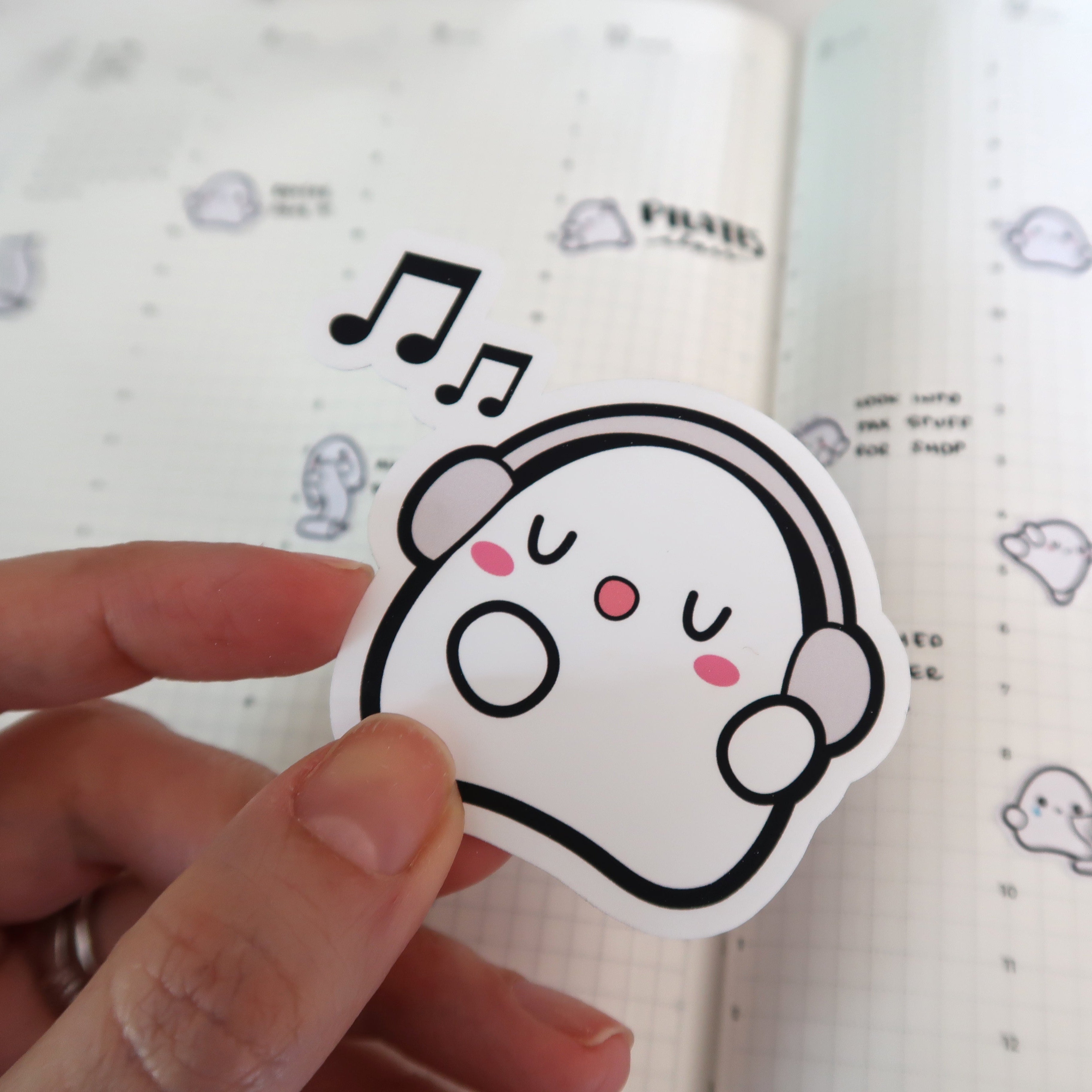 Sticker of a white cartoon ghost wearing grey headphones, singing happily with pink cheeks and black musical notes floating above, held by a hand against a grid-lined planner