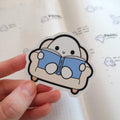 Blobbyland sticker with a smiling white blob character on a beige couch reading a blue book titled Blobbyland held by a hand over a light gridded notebook