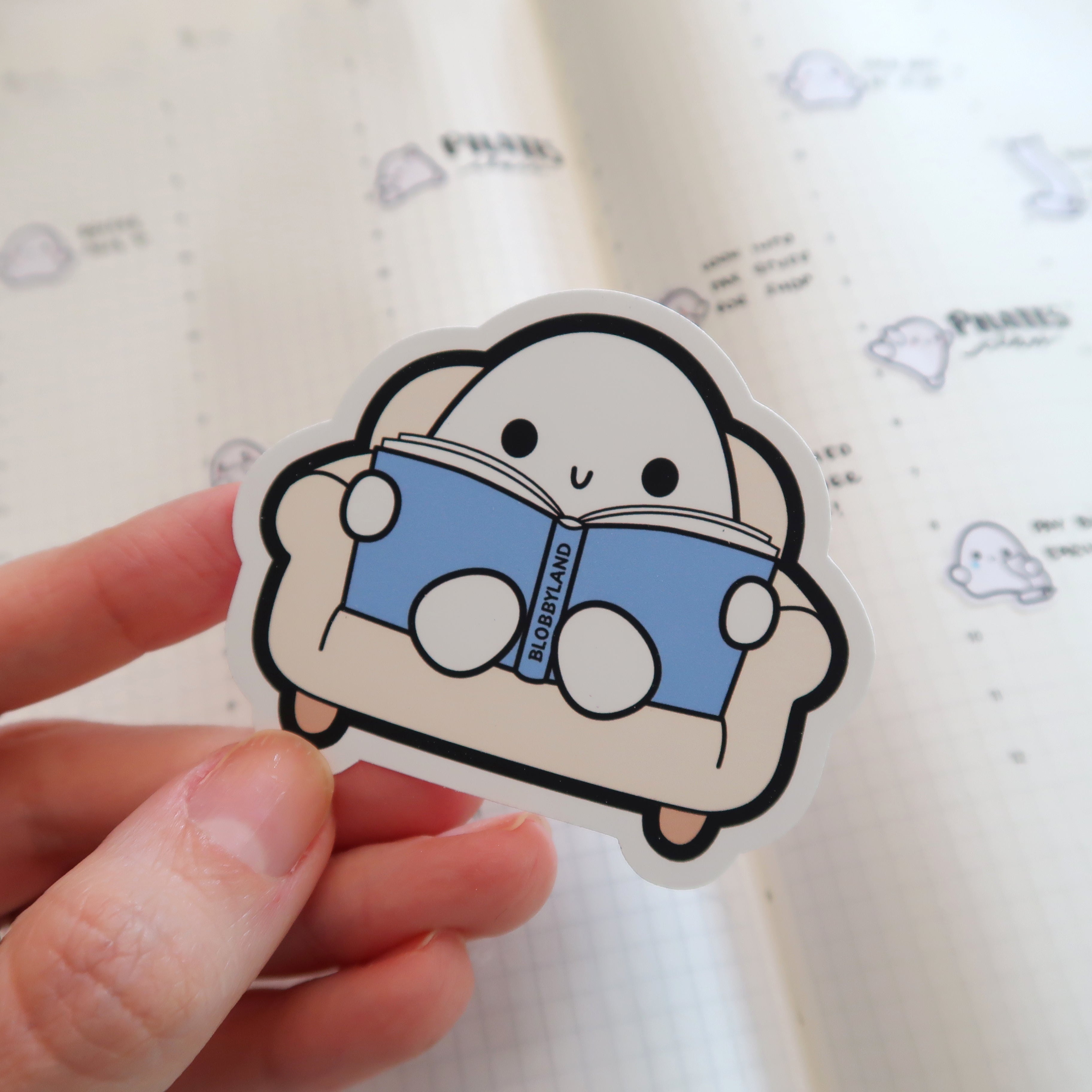 Blobbyland sticker with a smiling white blob character on a beige couch reading a blue book titled Blobbyland held by a hand over a light gridded notebook