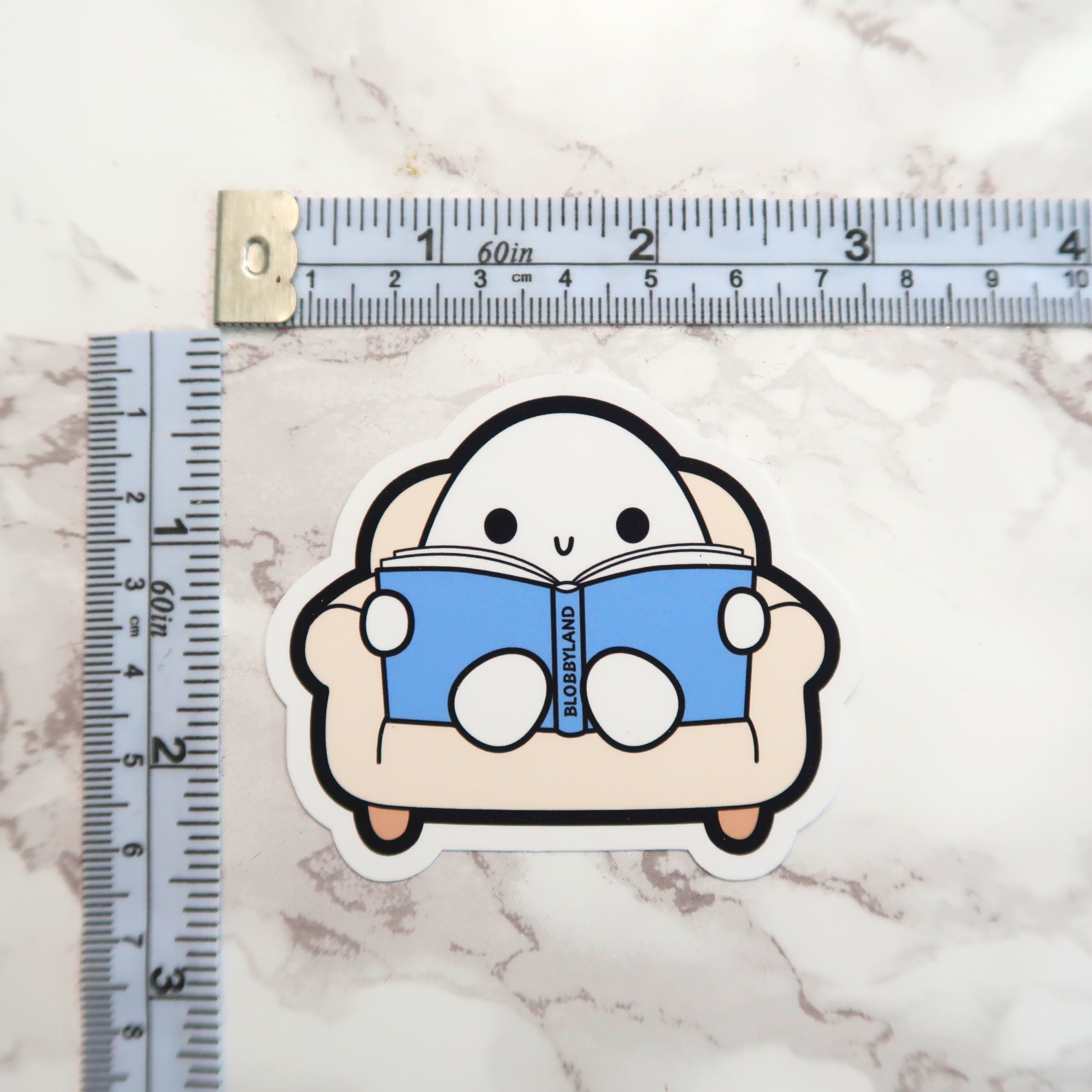 Vinyl sticker of a cute white blob character with black eyes and a happy U-shaped mouth sitting in a beige armchair, holding an open blue book titled 'BLOBBYLAND' on a light marble surface next to a white and blue measuring tape