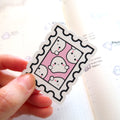A hand holds a playful postage stamp sticker featuring five white cartoon ghosts with black eyes and pink blush on a vibrant pink background above a blurred white planner with subtle cloud icons