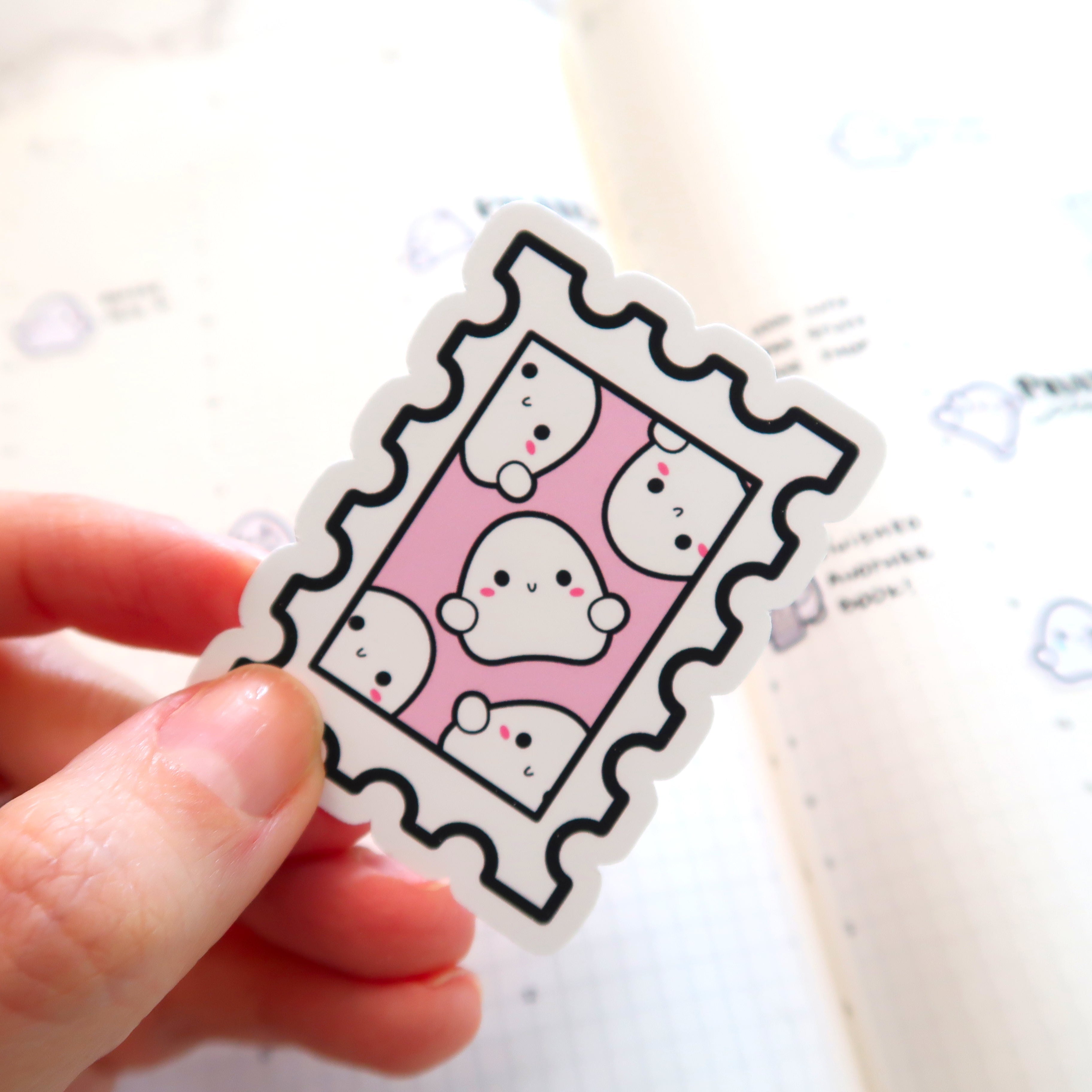 A hand holds a playful postage stamp sticker featuring five white cartoon ghosts with black eyes and pink blush on a vibrant pink background above a blurred white planner with subtle cloud icons