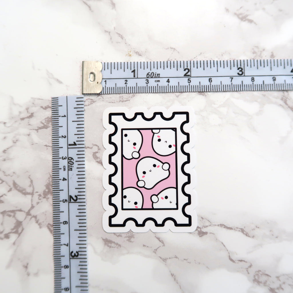 Die-cut sticker featuring a stamp design with six cute white cartoon ghosts showing rosy pink cheeks and black dot eyes on a pale pink background, placed on a white marble surface with a measuring tape visible