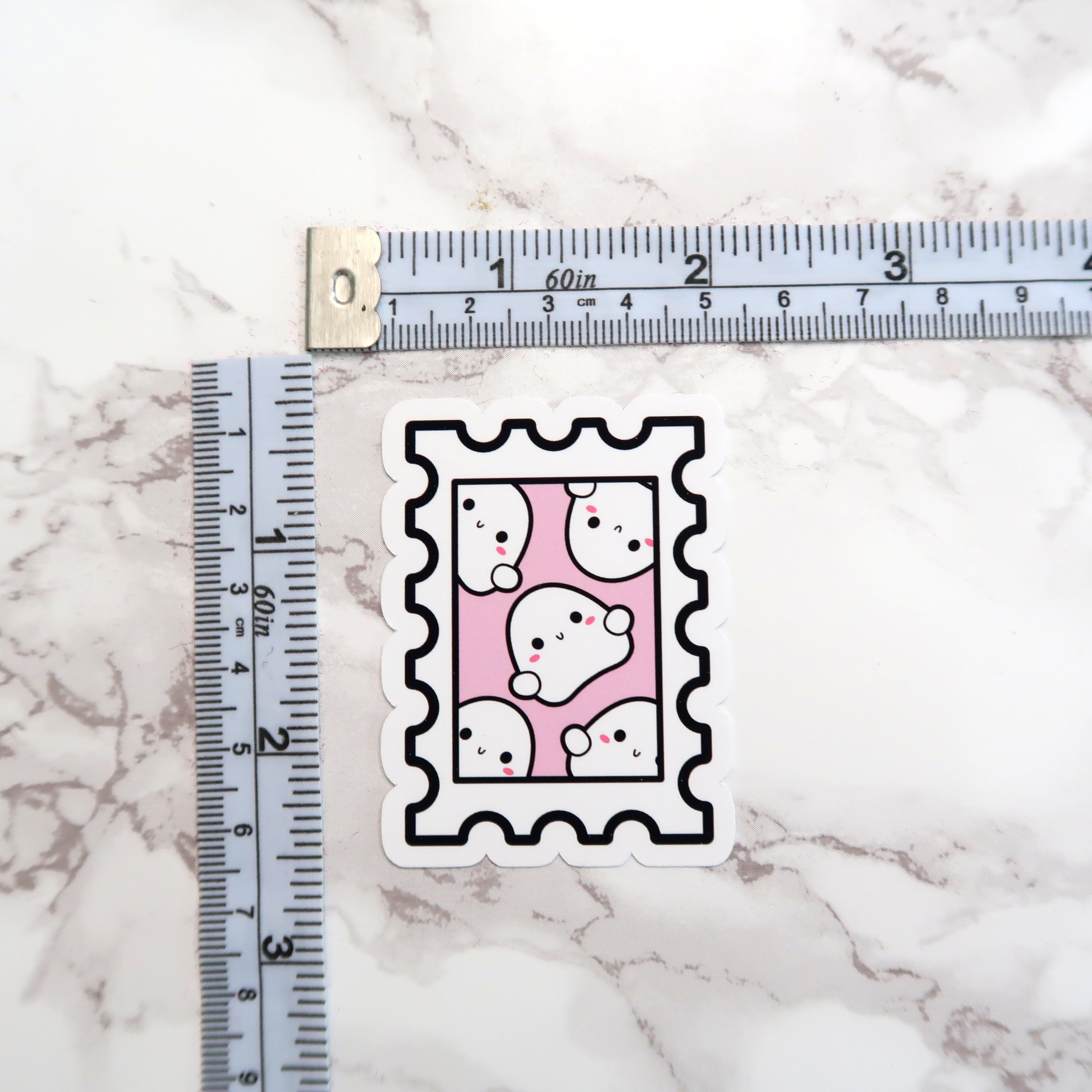 Die-cut sticker featuring a stamp design with six cute white cartoon ghosts showing rosy pink cheeks and black dot eyes on a pale pink background, placed on a white marble surface with a measuring tape visible