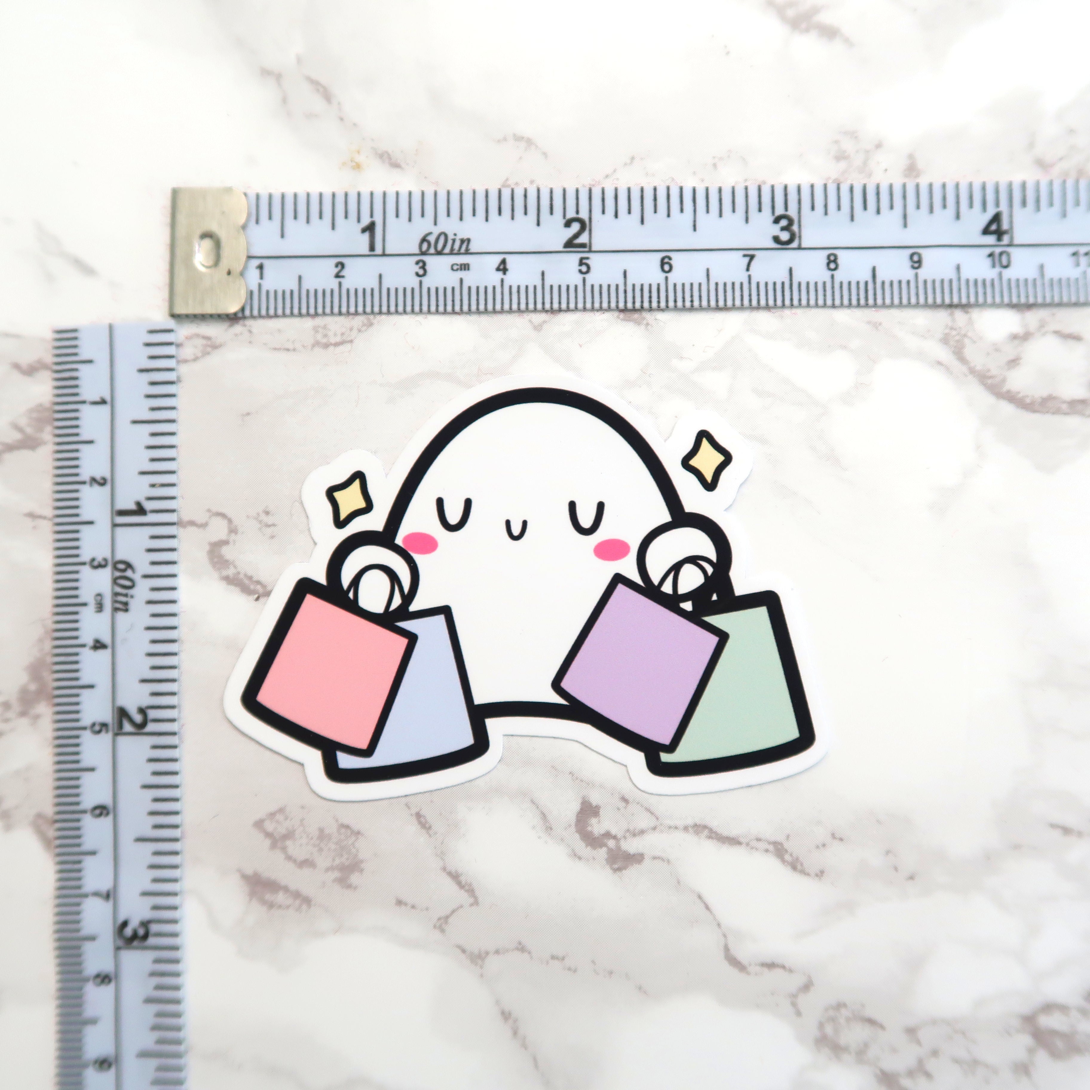 Sticker illustration of a happy white cartoon ghost with pink cheeks and yellow sparkles holding four pastel shopping bags (pink, light blue, lavender, light green) displayed on a white marble surface alongside a measuring tape with 60in and centimeter markings
