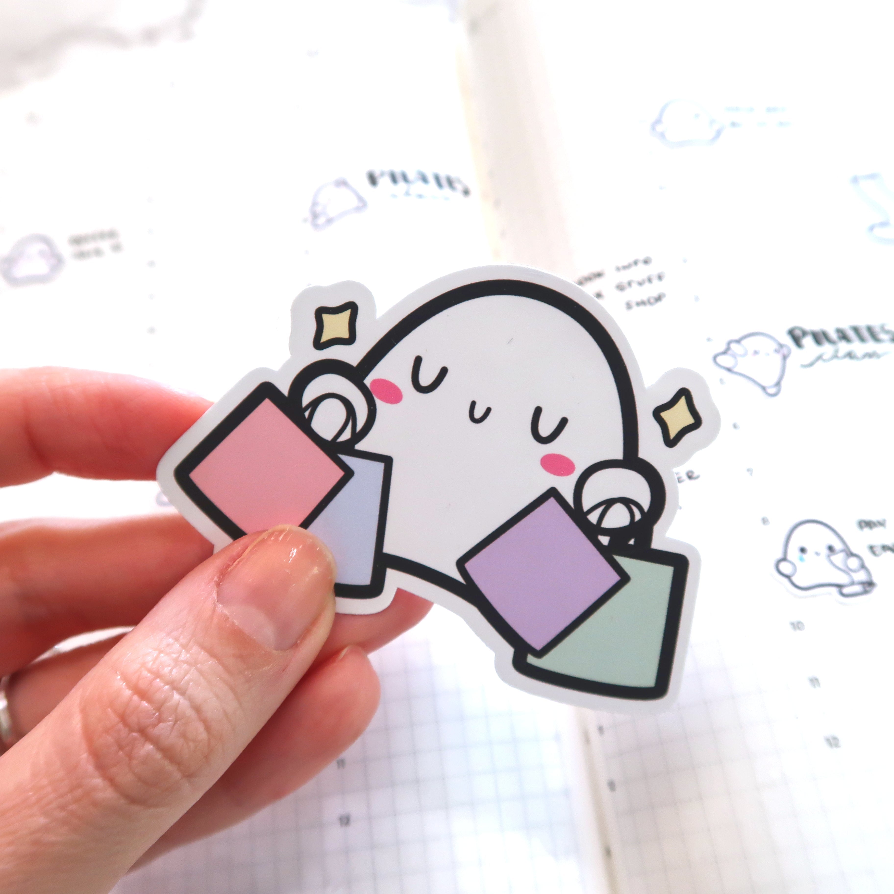 Die-cut sticker of a cute white ghost character with pink blush cheeks and sparkling yellow stars, holding pastel pink, purple, and mint green shopping bags, held by a hand over a bullet journal page showing "Pilates" and small ghost doodles