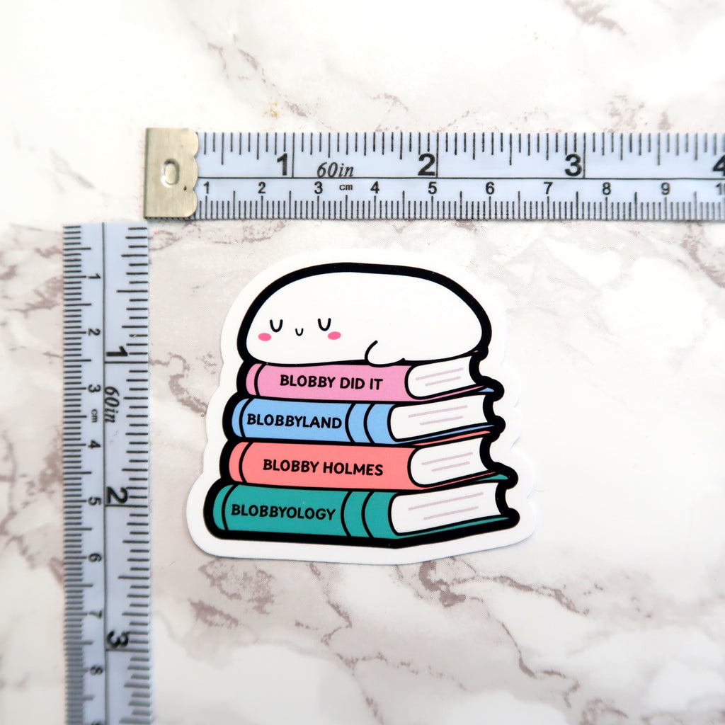 Sticker depicting a sleepy white blob character with pink cheeks and U-shaped eyes resting on a stack of pastel books titled Blobby Did It, Blobbyland, Blobby Holmes, and Blobbyology, positioned on a light marble surface beside a white and black measuring tape for scale