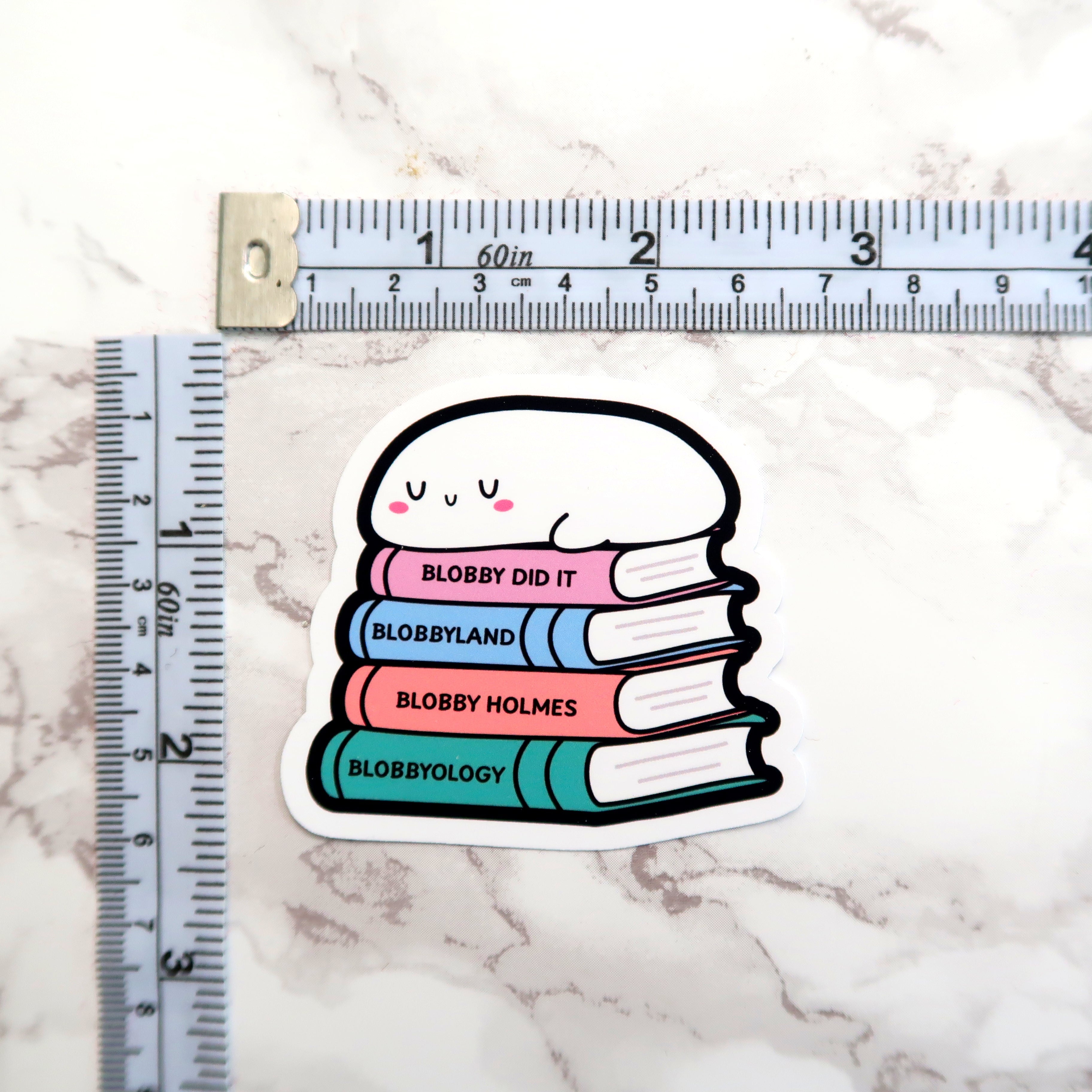 Sticker depicting a sleepy white blob character with pink cheeks and U-shaped eyes resting on a stack of pastel books titled Blobby Did It, Blobbyland, Blobby Holmes, and Blobbyology, positioned on a light marble surface beside a white and black measuring tape for scale