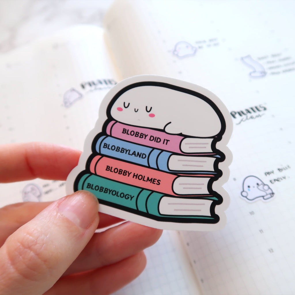 Photograph of a sticker showing a cute white blob character with pink cheeks and 'u' shaped eyes sleeping on a stack of pastel-colored books titled 'Blobby Did It', 'Blobbyland', 'Blobby Holmes', and 'Blobbyology' held by a hand over an open planner with grid lines and 'Pilates plan' written on it alongside other small blob doodles