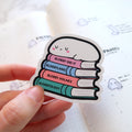 Photograph of a sticker showing a cute white blob character with pink cheeks and 'u' shaped eyes sleeping on a stack of pastel-colored books titled 'Blobby Did It', 'Blobbyland', 'Blobby Holmes', and 'Blobbyology' held by a hand over an open planner with grid lines and 'Pilates plan' written on it alongside other small blob doodles