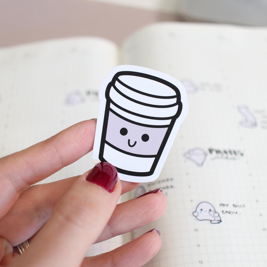 Hand holding a cute coffee cup sticker over a notebook with cartoon cloud designs.