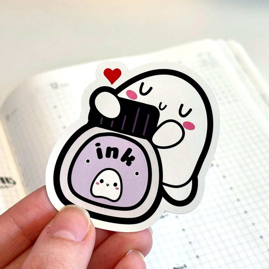 Hand holding a cartoon-style sticker of an ink bottle with a character on a notebook background