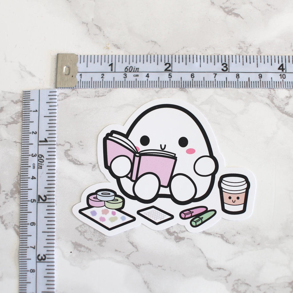 Sticker depicting a kawaii white egg character with pink cheeks reading a pink book, accompanied by a smiling coffee cup, colorful washi tape rolls, a pastel dot sticker sheet, and pink and green erasers, all arranged on a white marble surface next to a tailor's measuring tape