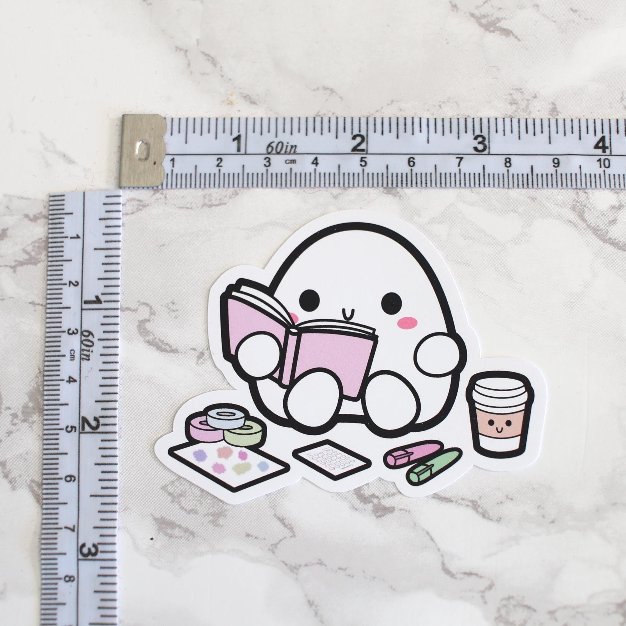 Sticker depicting a kawaii white egg character with pink cheeks reading a pink book, accompanied by a smiling coffee cup, colorful washi tape rolls, a pastel dot sticker sheet, and pink and green erasers, all arranged on a white marble surface next to a tailor's measuring tape