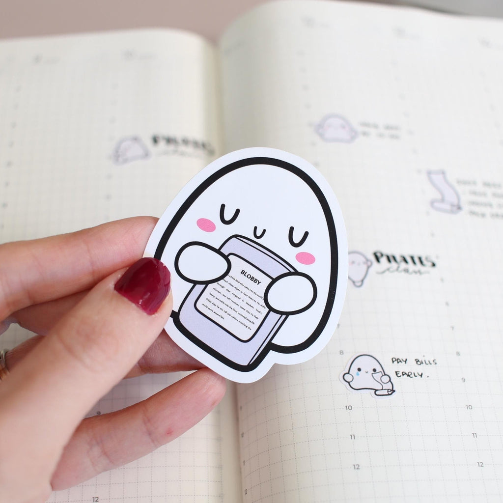 Hand holding a cartoon character sticker over an open notebook with more stickers on the page.